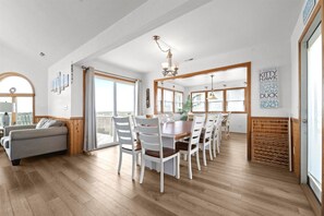 A spacious dining area with a wooden table and white chairs, large windows with a view, and a cozy seating area with a sofa and a lamp. - 8BR Oceanfront home - Game & theater rooms, private pool, hot tub ★★★★★ (Waves)
