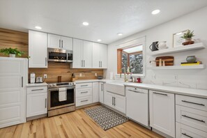 A modern kitchen with white cabinets, stainless steel appliances, a wooden backsplash, and a window with a view of trees.
