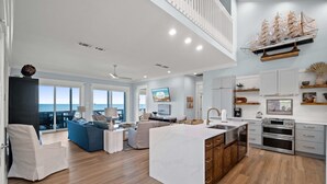 Un vaste espace de vie avec une grande fenêtre donnant sur la mer, une cuisine équipée d’appareils modernes et un coin repas agrémenté d’un canapé bleu.