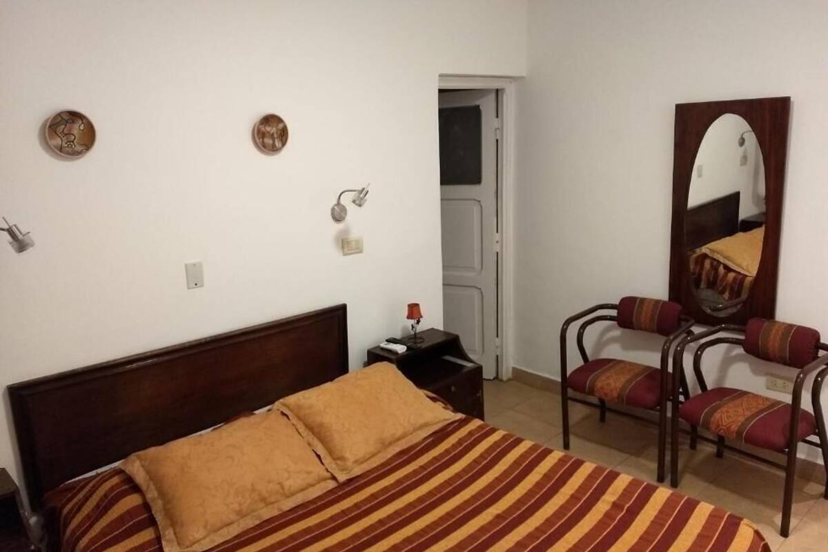 Quarto com cama, mesa de cabeceira, abajur, espelho e duas cadeiras.