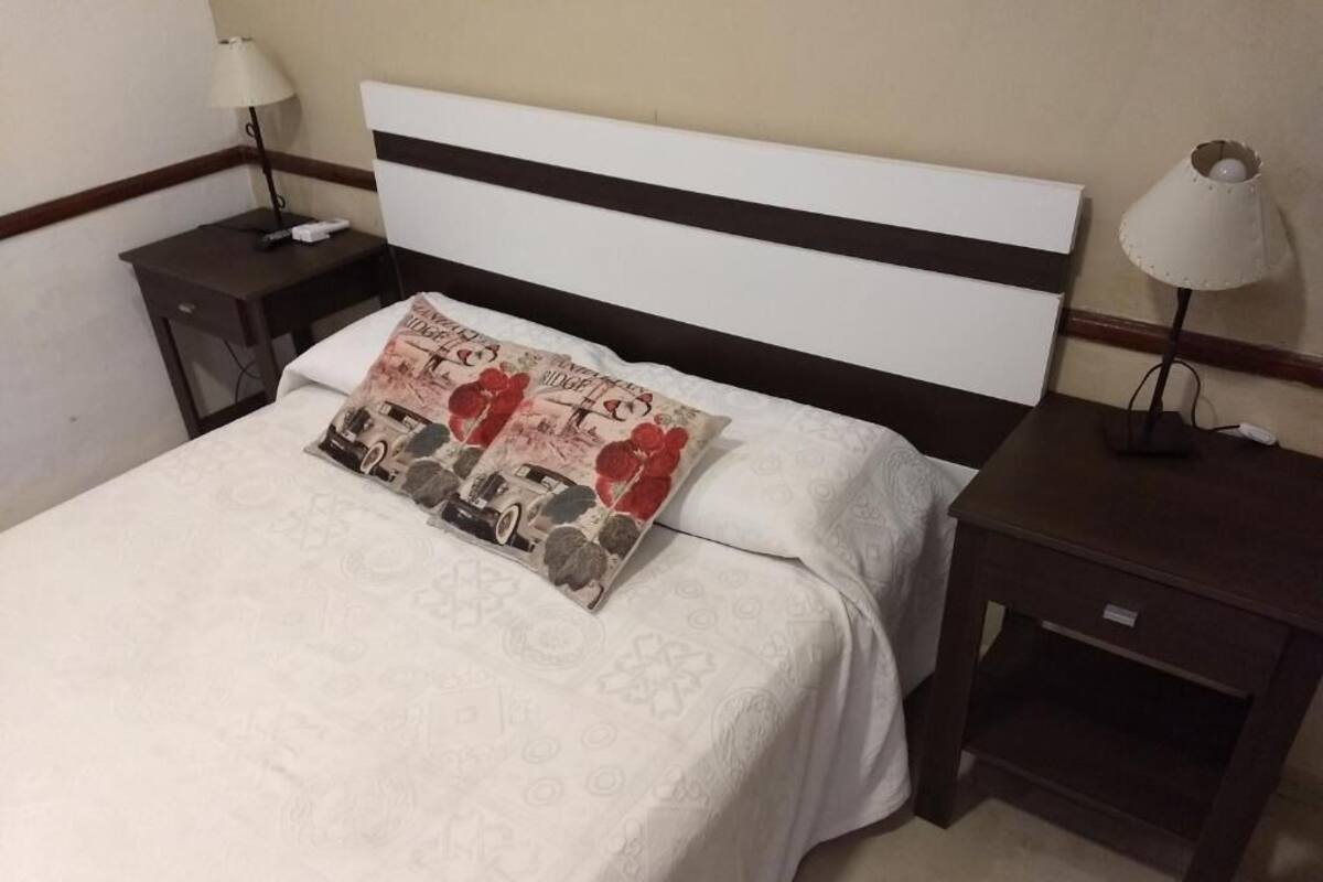 Cama bem arrumada com colcha estampada e almofada decorativa, com duas mesas de cabeceira com abajures de cada lado.