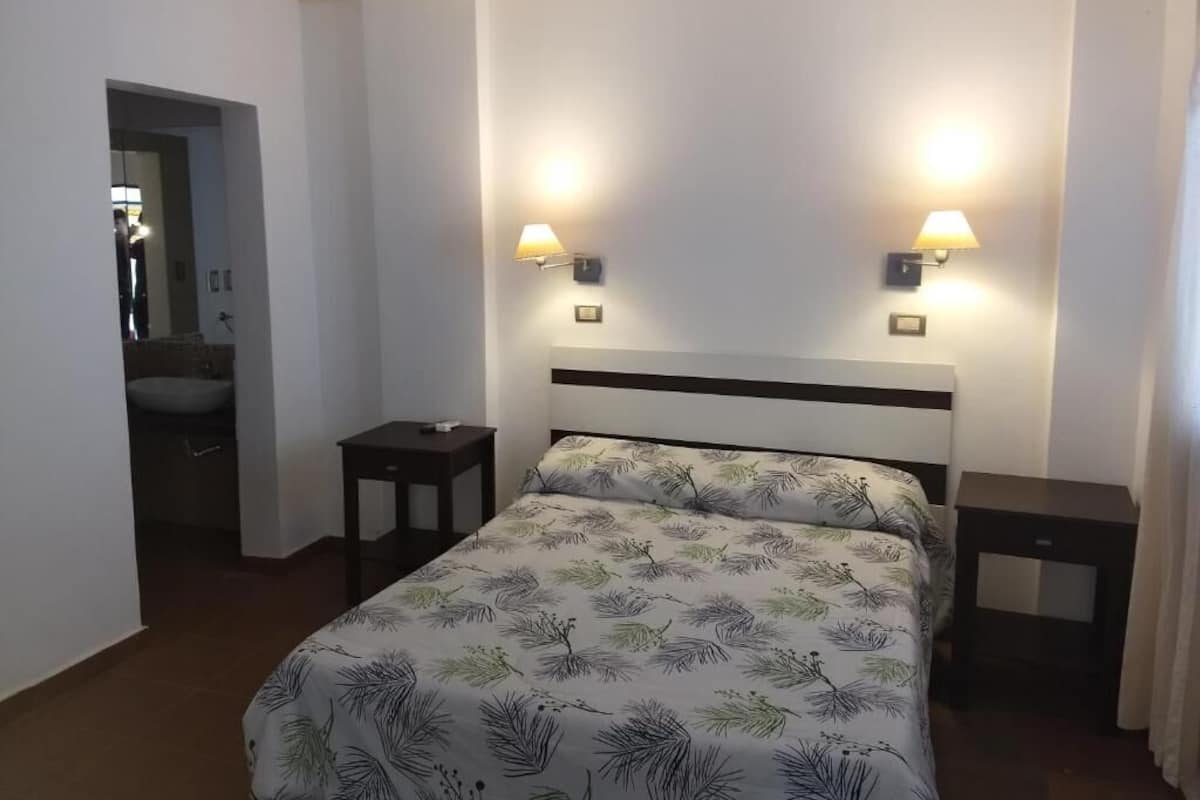 Quarto de hotel com cama, duas mesas de cabeceira, um criado-mudo, uma luminária na parede e uma porta que dá para o banheiro.