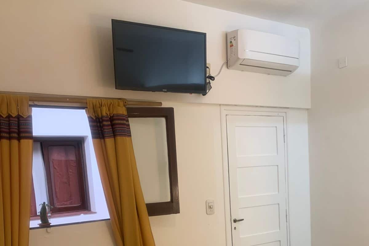 Quarto com cama, poltrona, televisão, ar condicionado, janela com cortinas e porta.