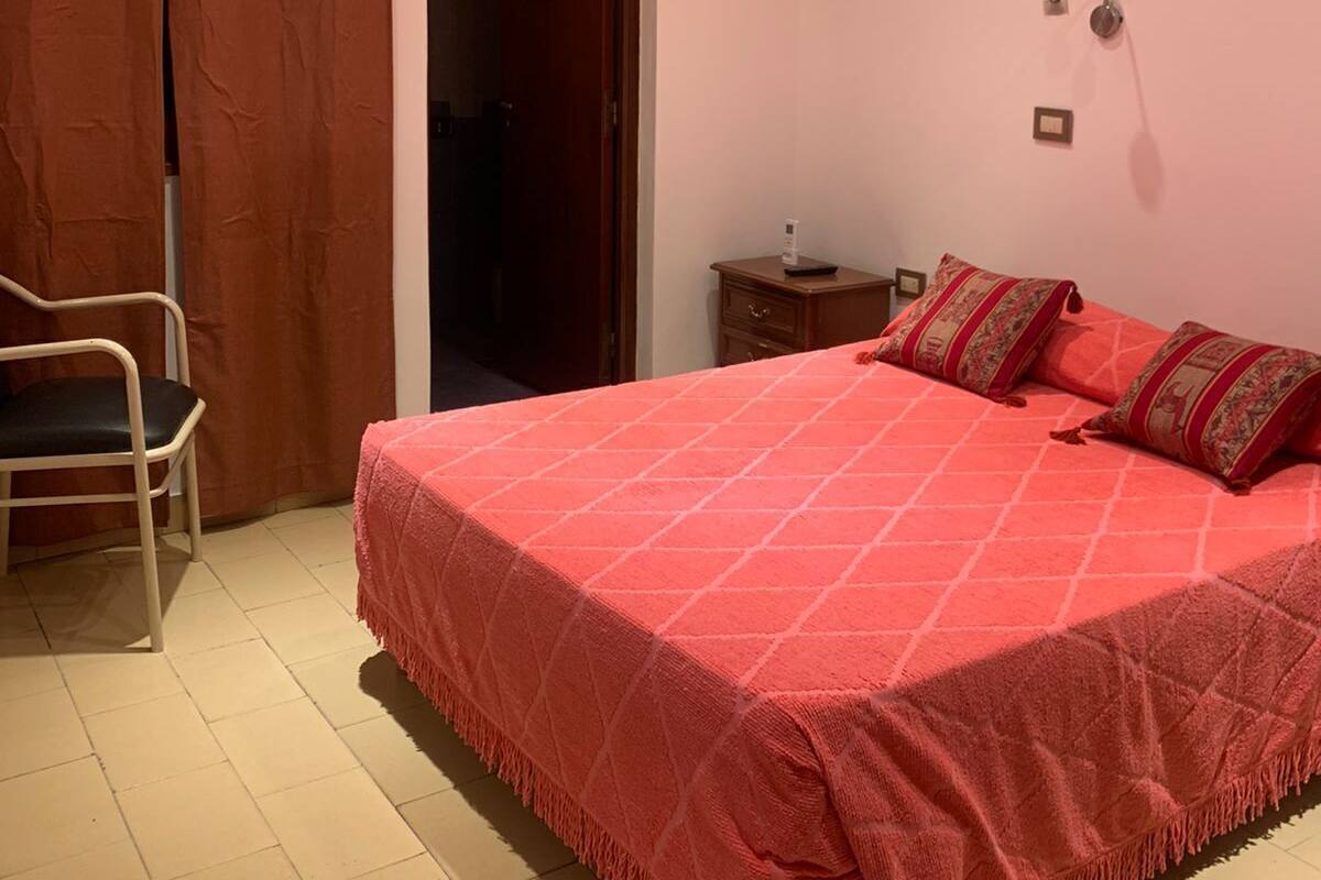 Quarto com cama, cadeira, mesa de cabeceira, luminária de parede e ar condicionado.