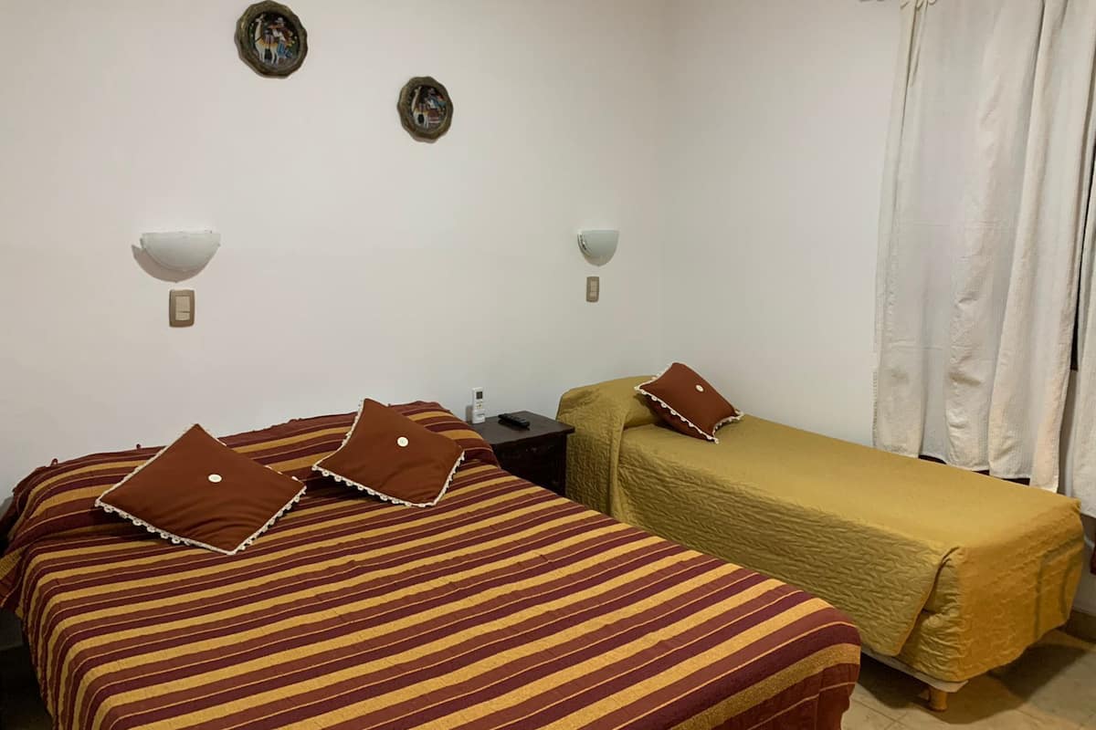 Quarto com duas camas, cada uma com roupa de cama e almofadas listradas, uma mesinha com abajur e cortina na janela.