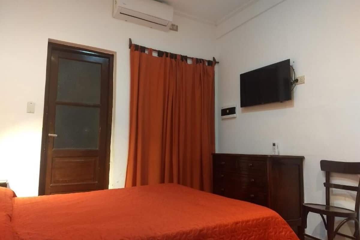 Quarto com cama, televisão na parede, penteadeira de madeira e porta com janela.