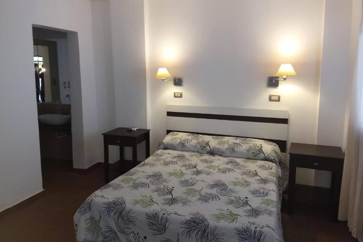 Quarto de hotel com uma cama, duas mesas de cabeceira, um criado-mudo, uma luminária na parede e uma porta para o banheiro.