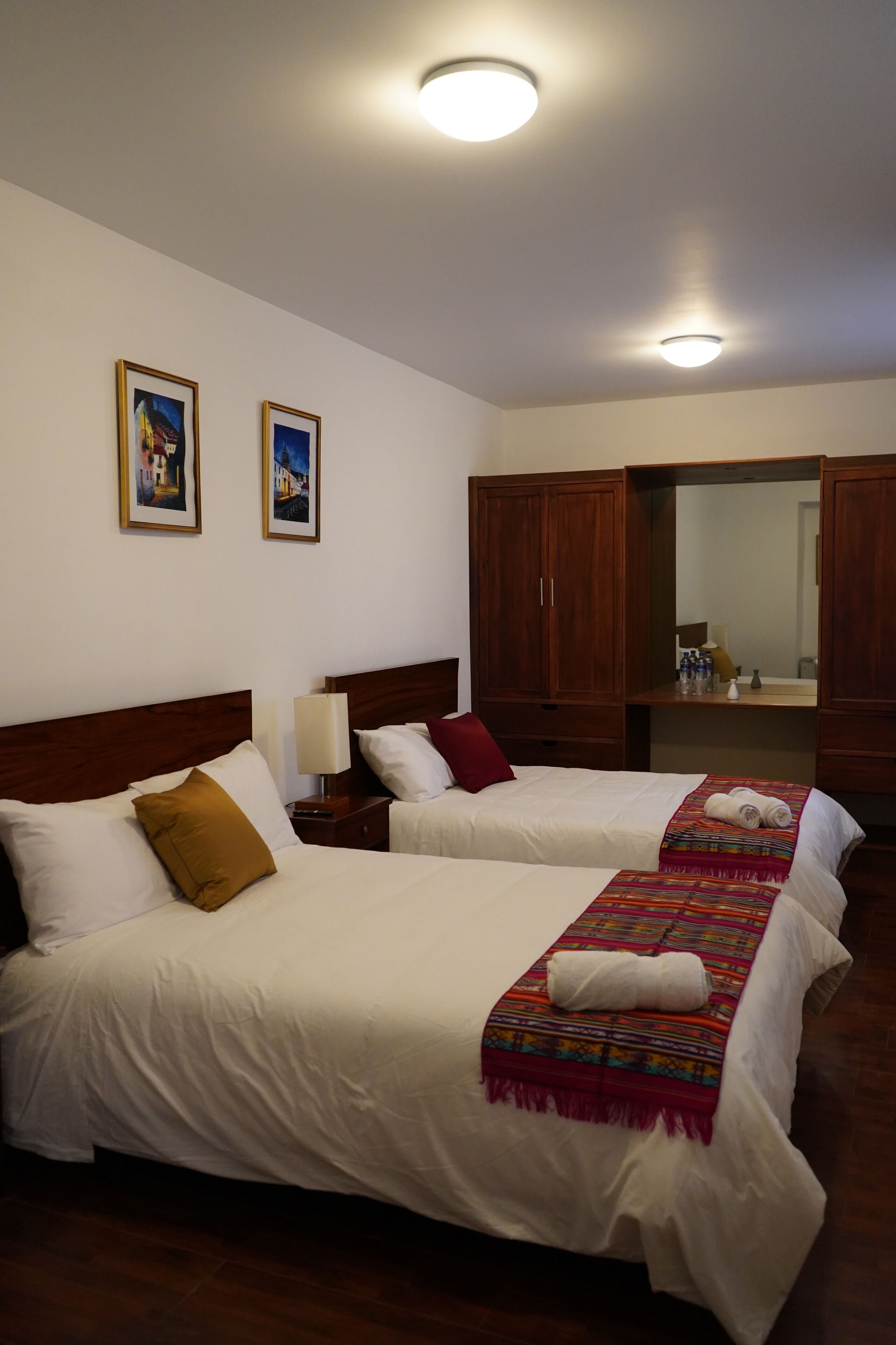 Habitación de hotel con dos camas, cabeceros de madera, ropa de cama blanca, un espejo y cuadros en la pared.