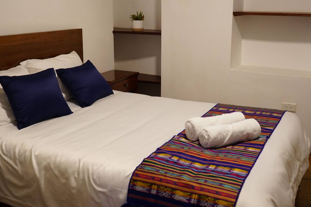A neatly made bed with white linens, two dark blue pillows, and a colorful woven blanket. There is a small potted plant on a shelf above the bed.