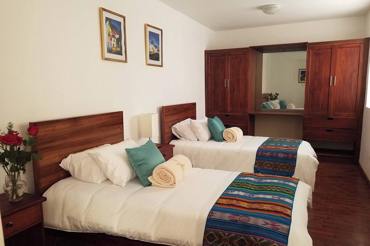 A hotel room with two beds, wooden headboards, white bedding, a colorful blanket, a vase with red roses, and two framed pictures on the wall.