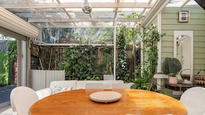 A covered patio with a wooden table and white chairs, surrounded by lush greenery and a white fence. - The Chelsea Cottage, Alfresco Living East Redfern (Redfern)