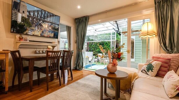 A cozy living room with a dining area, a large window with curtains, and a view of a garden. - The Chelsea Cottage, Alfresco Living East Redfern (Redfern)