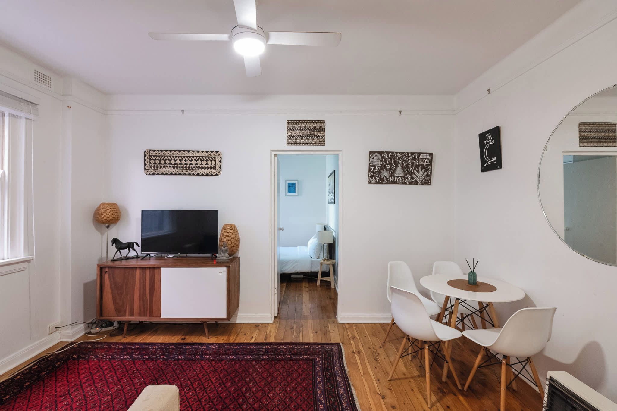 Ein modernes Wohnzimmer mit Flachbildfernseher, einer Holzkabine, einem roten Teppich mit Muster, weißen Stühlen und einem Deckenventilator.