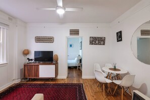 A modern living room with a flat-screen TV, a wooden cabinet, a red patterned rug, white chairs, and a ceiling fan.