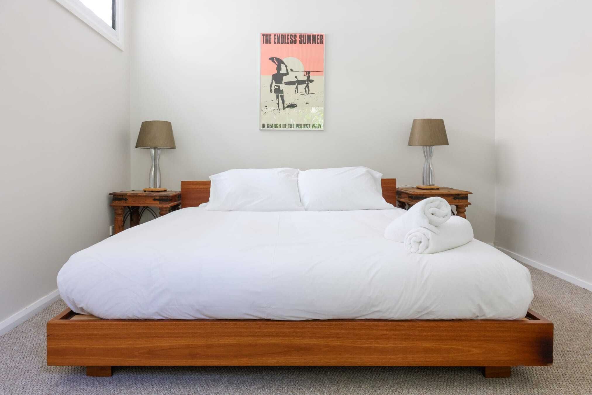 Una cama bien hecha con sábanas blancas y una toalla enrollada, dos lámparas de noche y un cartel en la pared.