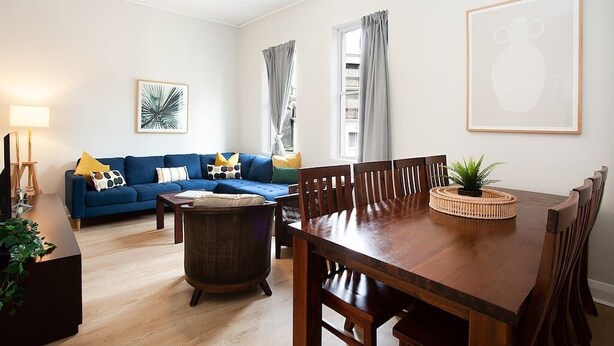 A living room with a blue sofa, a wooden dining table, and a coffee table. - Incredible Beachside Location 4 Bed Apartment (Bondi Beach)