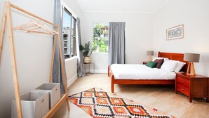 A bedroom with a wooden bed, bedside tables, a hanging clothes rack, and a view of greenery outside. - Incredible Beachside Location 4 Bed Apartment (Bondi Beach)