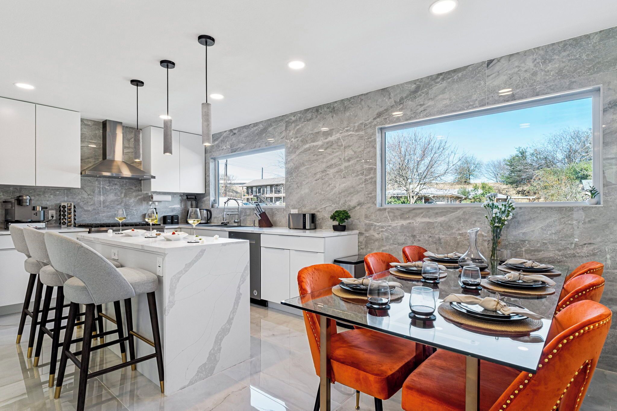 A modern kitchen with a central island, bar stools, and a dining area with a glass table and orange chairs.