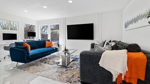 Een moderne woonkamer met een blauwe bank, een grijze bank, een glazen salontafel en een grote flatscreen televisie aan de muur.