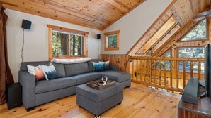 A cozy cabin interior with a wooden ceiling, a gray sectional sofa, a small ottoman, and a wooden deck with a railing. - Perfect chalet accommodations tucked into the most wonderful towering trees! (Deming)
