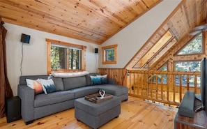A cozy cabin interior with a wooden ceiling, a gray sectional sofa, a small ottoman, and a wooden deck with a railing.
