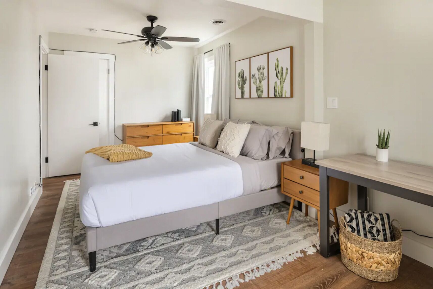 A bedroom with a bed, bedside tables, a desk, a ceiling fan, and a wall with cactus artwork.