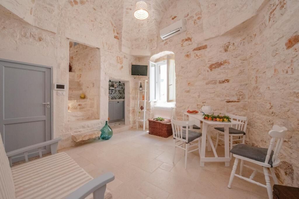 Un interior rústico con paredes de ladrillo visto, una zona de comedor con sillas blancas, una pequeña cocina y una puerta gris.