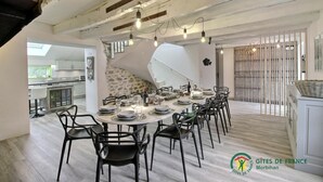 A spacious dining area with a high ceiling and exposed beams, a kitchen in the background, and a staircase leading to another level. - Heol - AMBON - Gîte (AMBON)