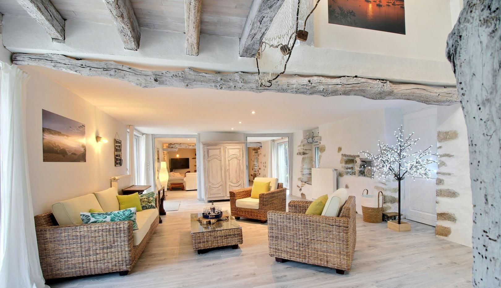 Un vaste espace de vie aménagé avec des meubles en osier, une table basse et un arbre décoratif. On aperçoit une autre pièce avec un lit et une armoire.