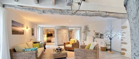 Amplia sala de estar con muebles de mimbre, una mesa de centro y una planta decorativa. Se ve una habitación con una cama y un armario.