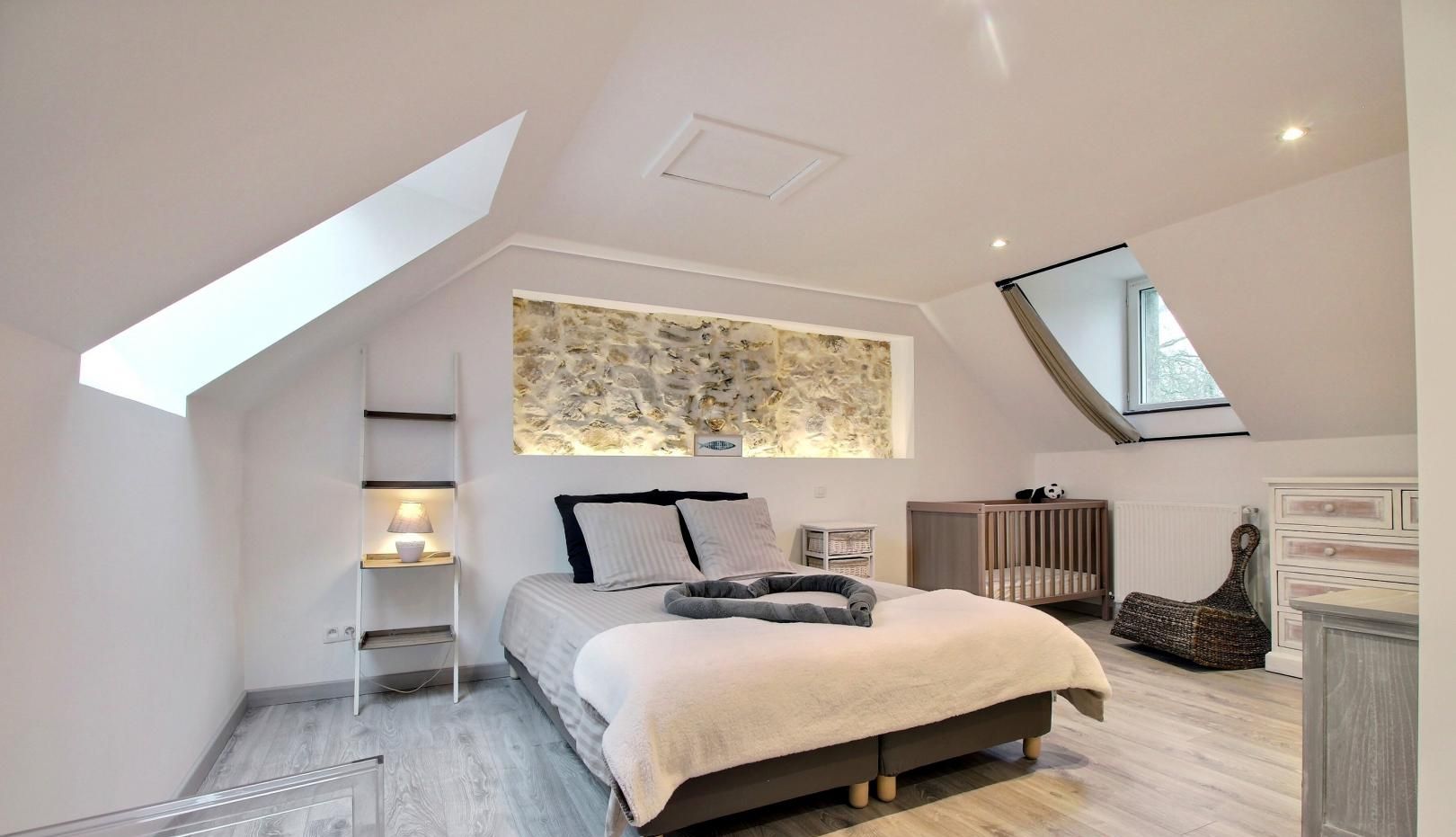 Un dormitorio con techo inclinado, una cama con cabecera, una mesita de noche, una cuna, un aparador y una obra de arte colgada en la pared.