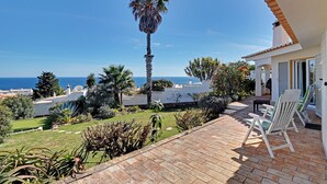 A patio with a view of the sea, a palm tree, and a white building with a tiled roof.