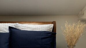 Una cama bien hecha con ropa de cama blanca y azul marino, un cabecero de madera y un pequeño taburete de madera con un jarrón de plantas secas a su lado.