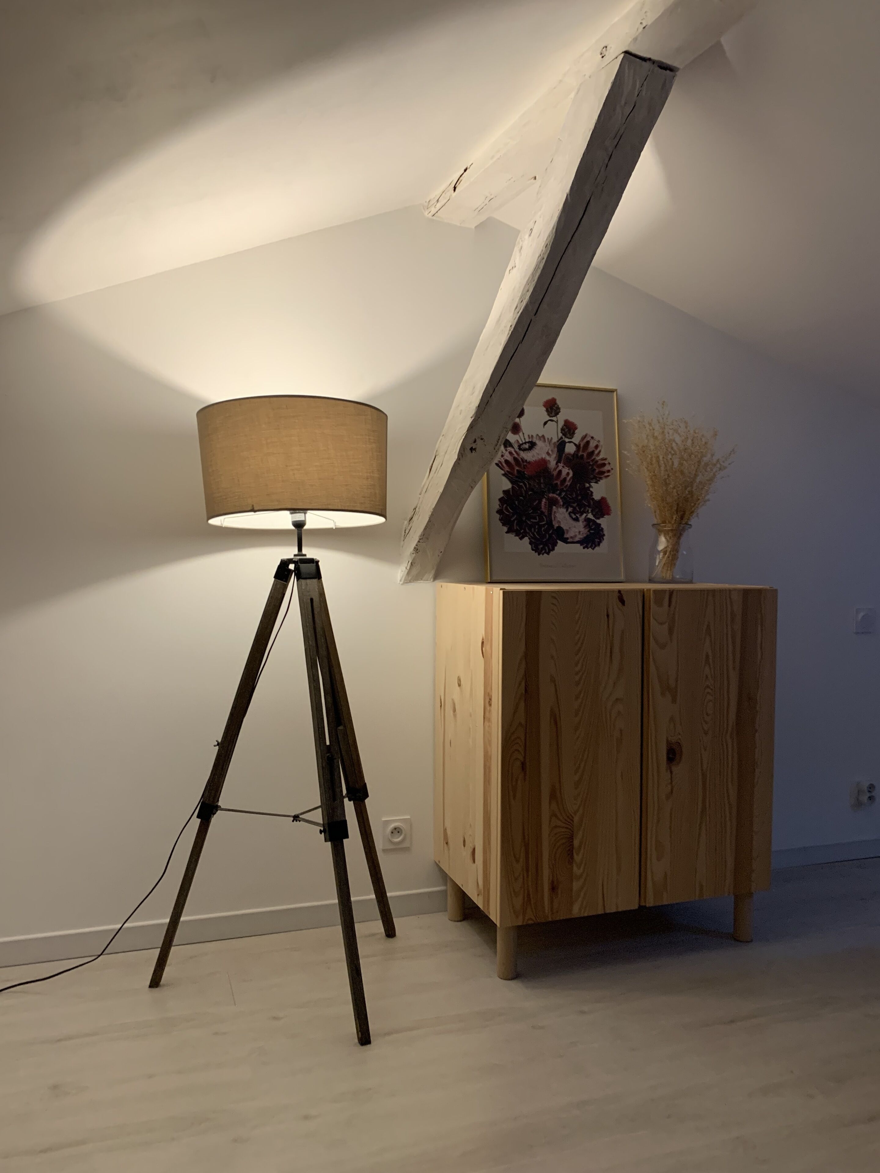 Et værelse med trægulv, en tripod gulvlampe, et træskab og et indrammet blomsterbillede.