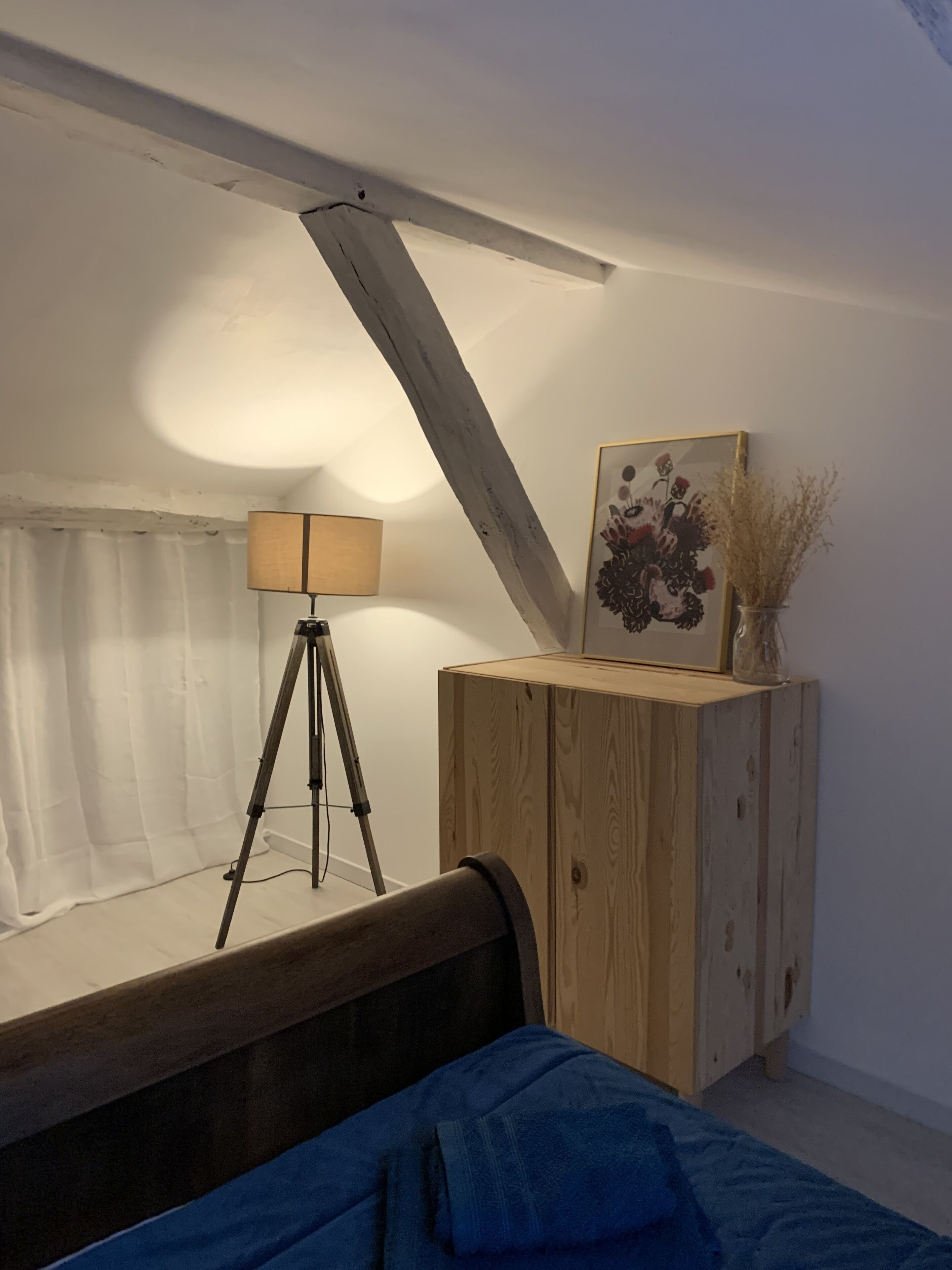 A bedroom with a wooden headboard, a tripod with a lamp, a wooden cabinet with a vase, and a framed picture on the wall.