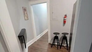A narrow hallway with wooden flooring, white walls, and a white door at the end. There are two black stools against the wall and a fire extinguisher mounted on the wall. - In Through The Side Door (Victorian District) (Savannah)