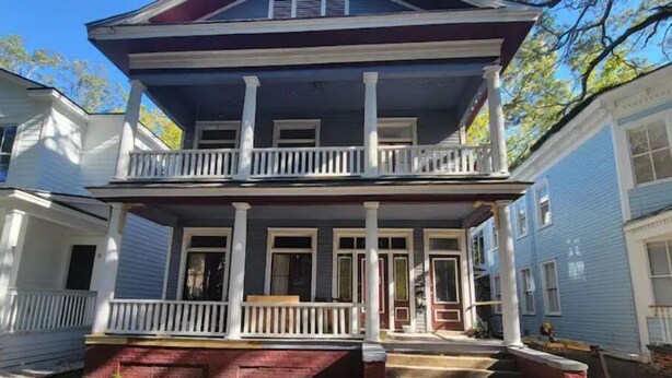 A two-story house with a front porch, a red brick foundation, and a staircase leading to the entrance. - In Through The Side Door (Victorian District) (Savannah)