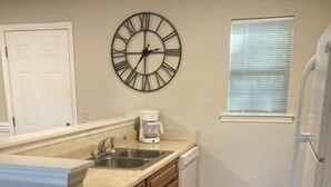 Une cuisine avec un plancher en bois, une horloge au mur et une fenêtre avec des stores.