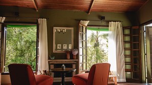 A room with two red chairs, a small table, a mirror, and a view of greenery through open windows. - Reserva Mata Grande - Nature Park (Biritiba-Mirim)
