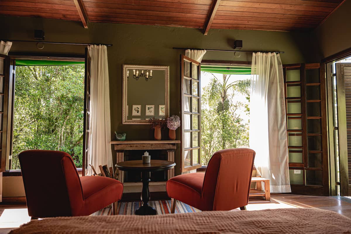 Ein Zimmer mit zwei roten Stühlen, einem kleinen Tisch, einem Spiegel und Blick auf Grünpflanzen durch offene Fenster.
