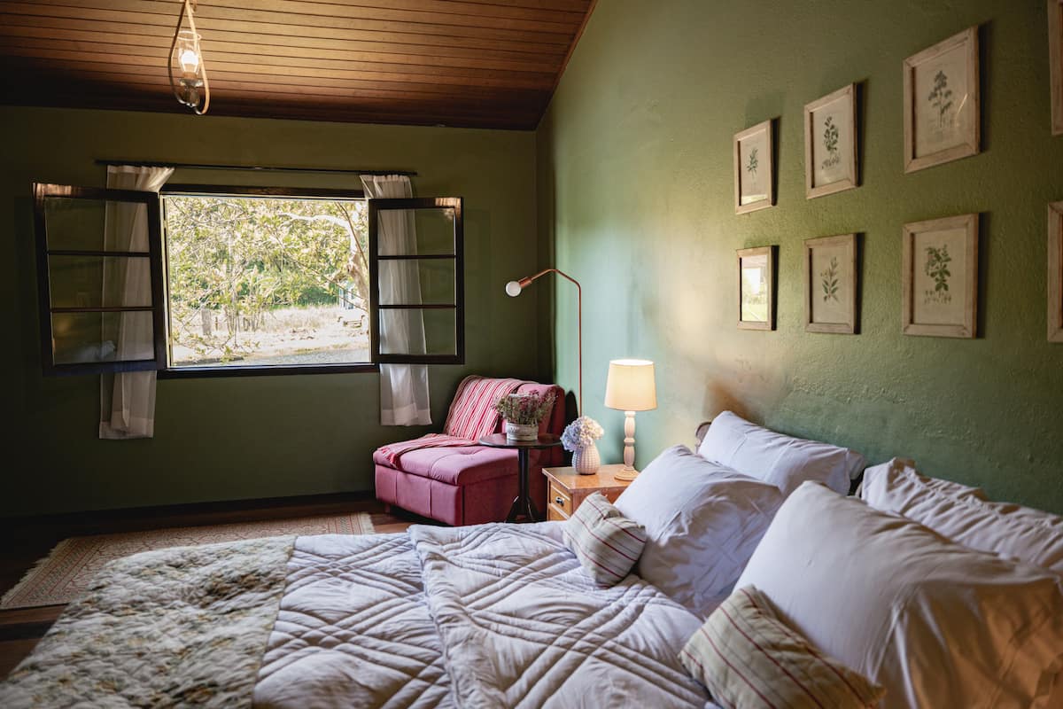Ein Schlafzimmer mit einem großen Bett, einem Fenster mit Vorhängen, einem Nachttisch mit Lampe sowie gerahmten Pflanzenbildern an der Wand.
