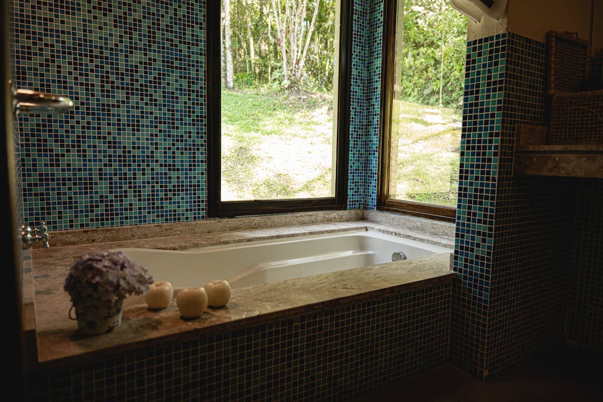 Ein Badezimmer mit Badewanne, einem Fenster mit Blick auf Grünflächen und einer gefliesten Wand.