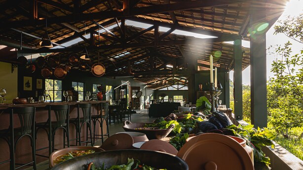 A restaurant with a rustic interior, featuring a high ceiling with exposed beams, hanging pots, and a variety of seating arrangements including bar stools and tables. - Reserva Mata Grande - Nature Park (Biritiba-Mirim)