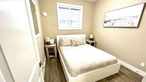 A bedroom with a bed, two bedside tables, a nightstand, a window with blinds, and a wall-mounted picture.