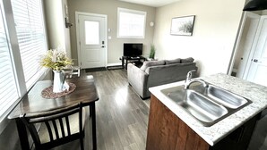 A modern kitchen with a built-in sink, wooden dining table, and chairs. There is a gray sofa in the living area and a small plant on the windowsill.