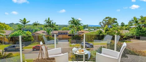 A balcony with wicker chairs, a table with a tray of food and drinks, and a view of a residential area with houses and palm trees.