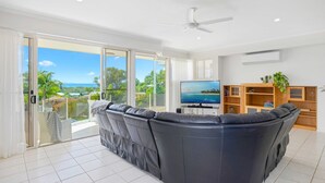 Una sala de estar moderna con un sofá seccional grande, un televisor de pantalla plana y vistas a un paisaje tropical a través de amplias puertas correderas de vidrio.