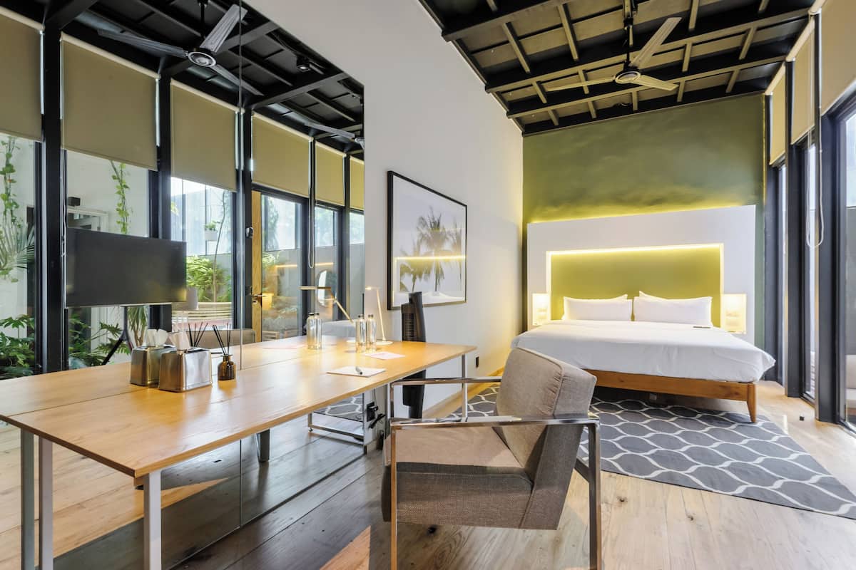 Quarto de hotel moderno com escrivaninha de madeira, uma cadeira confortável, uma cama com cabeceira grande e vista para o verde pelas janelas.