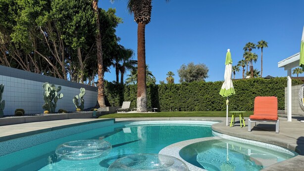 Una piscina con agua cristalina, rodeada de palmeras y un seto. Hay sillones de descanso y una pequeña mesa cerca.
