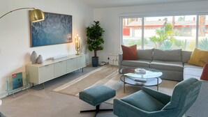 Una sala de estar moderna con un sofá, sillones, una mesa de centro de vidrio, una lámpara de pie, una planta y un cuadro en la pared.